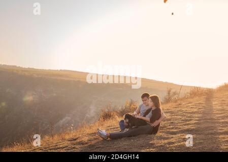 Sitting on a canyon,  looking at paragliders flying in the sky at sunset. Trip with pets. Young family with a schnauzer dog travel. Moldova, Old Orhey Stock Photo