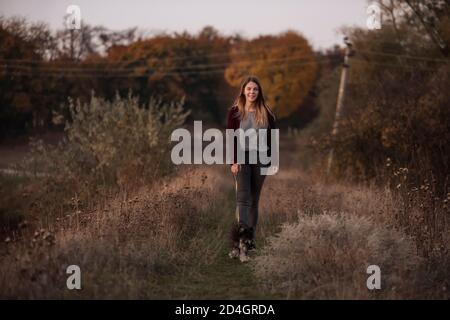 Modern Caucasian girl walks on a leash a Schnauzer dog in an autumn field at sunset. Dressed in dark jeans, burgundy cardigan. Taking care of pets. Stock Photo