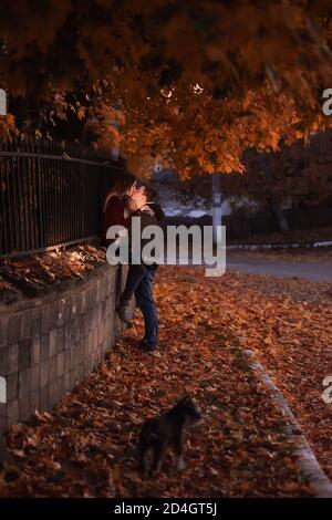 A young couple of teenagers are sitting by the fence under the orange foliage of a tree in the autumn evening. Lovers hug, look at each other, kiss. Stock Photo