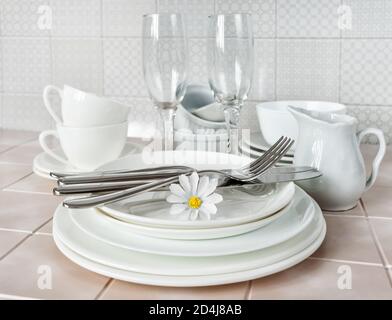 Stack of white porcelain plates, tea cups and wine glasses on the kitchen table against the background of a apron made of ceramic tile Stock Photo