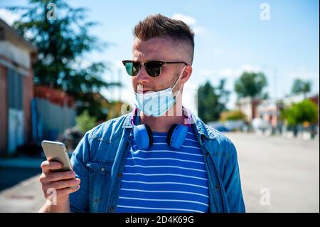 Cool guy wearing protective face mask standing outdoors while video calling his friends - Contemporary man having fun with phone during coronavirus ou Stock Photo