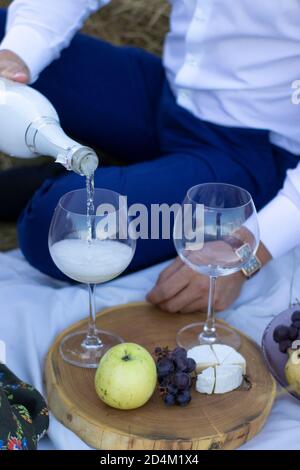 man's hand pouring champagne in glass on wooden stand with grapes and camembert cheese on a white blanket. picnic.  Stock Photo