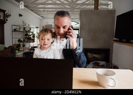Father talking on smartphone while watching videos on laptop with youngest daughter sitting in kithen. Stock Photo
