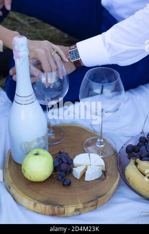 two hands holding champagne glasses near wooden stand with bottle, grapes and camembert cheese on white blanket. picnic. High quality photo Stock Photo