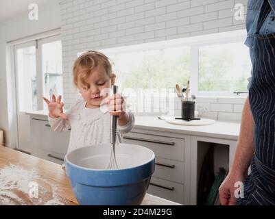 Baby girl whisking in a mixing bowl in the kitchen with father Stock Photo