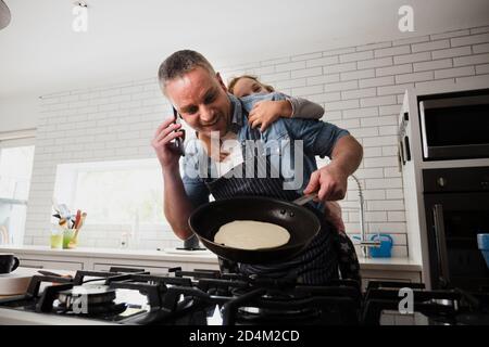 Father and daughter bonding together while baking pancakes chatting on smartphone in kitchen. Stock Photo