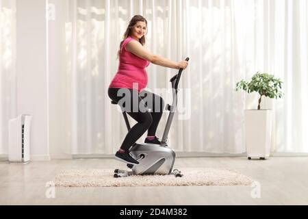 Pregnant woman in a pink top riding an exercise bike at home and looking at camera Stock Photo