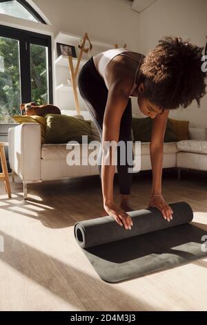 Beautiful African American female stretching before exercise routine holding gym mat in modern lounge. Stock Photo