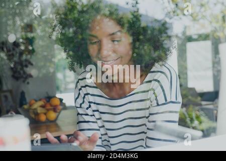 portrait of beautiful ethnic female scrolling on tablet smiling through window sill in casual kitchen. Stock Photo