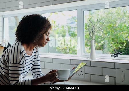 African American focused female reading online book off digital tablet while holding hot coffee standing in modern kitchen. Stock Photo