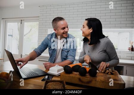 Happy couple smiling at each other following recipe on laptop while making healthy dinner in kitchen. Stock Photo