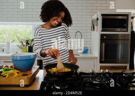 Smiling African American female making delicious dinner chatting to boyfriend on smartphone preparing for date night in fancy kitchen. Stock Photo