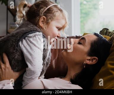 Mother giving young girl toddler a kiss while lying comfortably on sofa in living room. Stock Photo