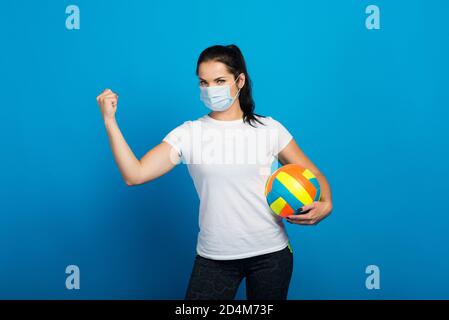 Happy sporty woman with face mask from coronavirus holding ball. Isolated on blue Stock Photo
