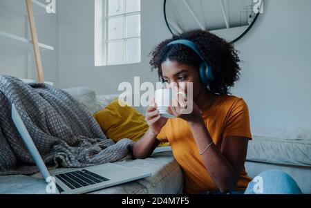 teenage student wearing headphones watching lectures on laptop drinking coffee sitting next to leather couch in trendy lounge. Stock Photo
