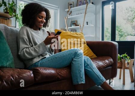 Beautiful young mixed race lady sitting on the couch at home scrolling through smartphone, smiling and happy Stock Photo