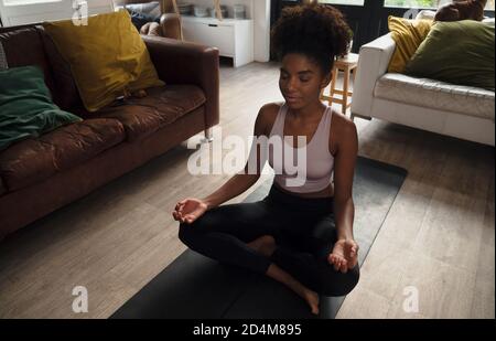 Young female calming meditating cross- legged with palms on knees smiling while sitting on gym mat in spacious lounge. Stock Photo
