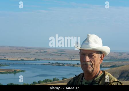 Portrait of senior man in hat. An elderly man stands on mountain, below there is a river and beautiful landscape. Seniors lifestyle concept. Stock Photo
