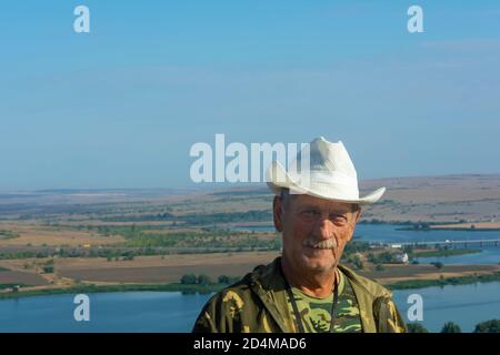 An older man in white straw hat looks at the camera against background of river and nature. Seniors lifestyle concept. Copy space, soft focus, blur. Stock Photo