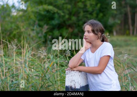 A beautiful teenage girl in white clothes in the forest looks to the side. Portrait in the park outdoors. Emotions, feelings concept. Stock Photo