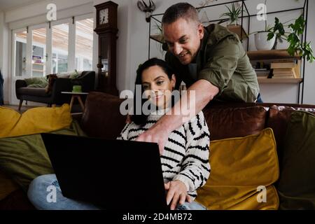 Handsome husband pointing at laptop while wife sits on comfy sofa in living room. Stock Photo