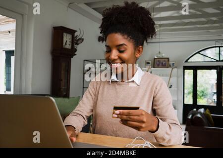 Happy African American business woman making online payment using fancy laptop sitting in modern kitchen. Stock Photo