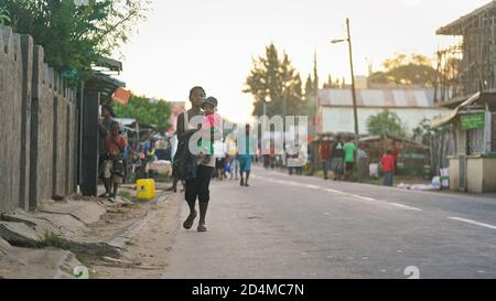 Ranohira, Madagascar - April 29, 2019:  Unknown Malagasy woman carrying baby kid, walking on asphalt road at small African city, more blurred people i Stock Photo