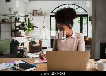 Mixed race female student studying online writing notes on notepad reading off laptop sitting in modern kitchen. Stock Photo