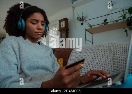 Mixed race female frowning while making online payment using laptop sitting on sofa in trendy lounge. Stock Photo