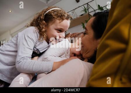 Mother and two daughters playing together and laughing while lying with each other on comfy sofa in living room Stock Photo