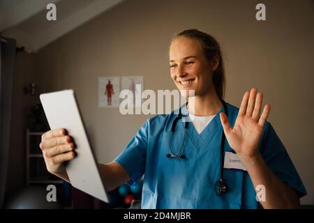 Beautiful young caucasian female doctor on video call from practice at home, smiling and waving at patient Stock Photo