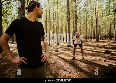 Healthy caucasian son waits for tired elderly father bending over knees in luscious woods.  Stock Photo