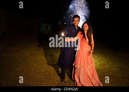 Young Indian couple in traditional wear celebrating holding sparklers, firecrackers or phuljhadi in hand as a part of diwali celebration, copy space t Stock Photo