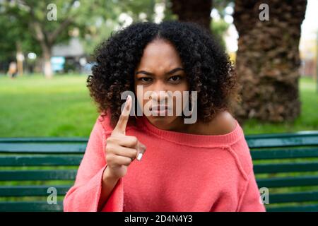 Portrait of young afro american woman with angry expression and pointing to camera while sitting on bench in the park. Outdoors. Stock Photo