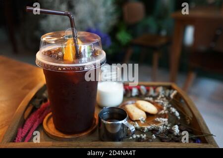 Close up plastic take away cup of iced black coffee served with milk, sugar syrup and butter cookies on wooden tray table decorated with dry flower Stock Photo