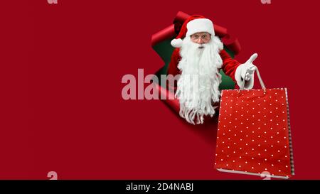 Santa man holds shops packet in hand through a paper hole. Bearded man in santa hat looking through hole on red paper. Stock Photo
