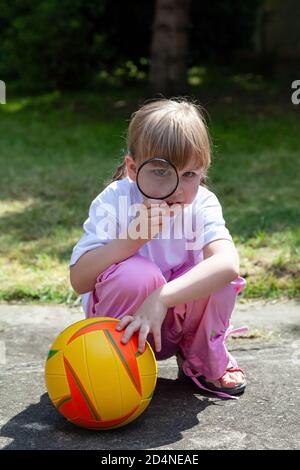 Small clever happy girl, child looking through a magnifying glass, kid with a loupe and a basketball playing in the garden outside. Curious little gir Stock Photo