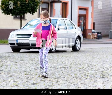 Lone little school age girl, little child going shopping in a protective face mask, holding a shopping list, walking alone on the street, new normal Stock Photo