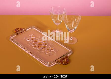 Two vintage classsic martini cocktail glasses standing next to a glass serving plate with flowers on a brown surface and a pink background. Clean mini Stock Photo