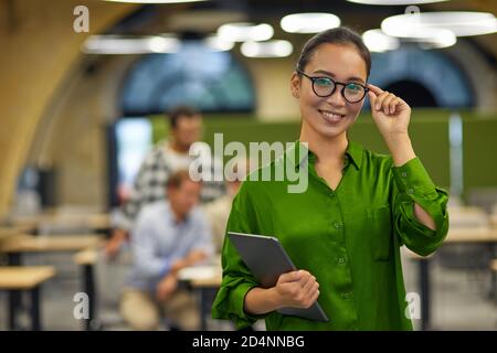 Portrait of young happy asian woman adjusting her eyeglasses, holding digital tablet and smiling at camera while standing in the office with colleagues on the background. Business people at work Stock Photo
