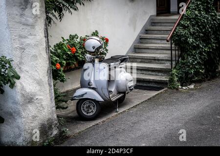 Lonely Vespa Scooter Iwrith a helmet on the bar in an Italian village in front of red flowers, stairs and a wall