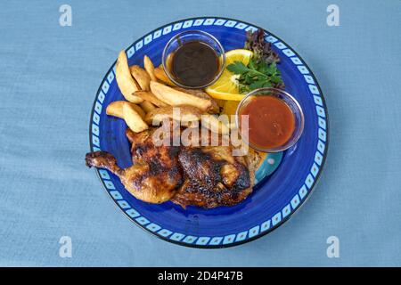 Grilled half chicken with potato wedges and two sauces on a rustic blue plate and tablecloth, copy space, high angle view from above, selected focus Stock Photo