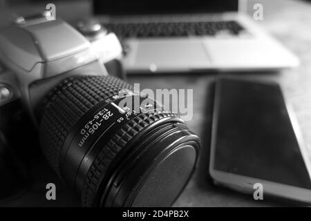 An old SLR 35mm camera placed on a coffee table next to a modern smartphone and laptop Stock Photo