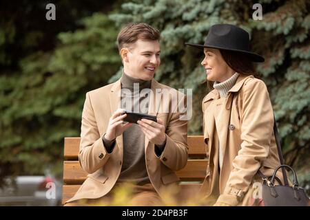 Happy young stylish man with smartphone and his pretty girlfriend having chat Stock Photo