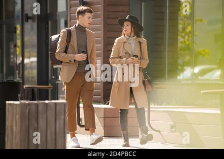 Young serious stylish woman with drink talking to her boyfriend in casualwear Stock Photo
