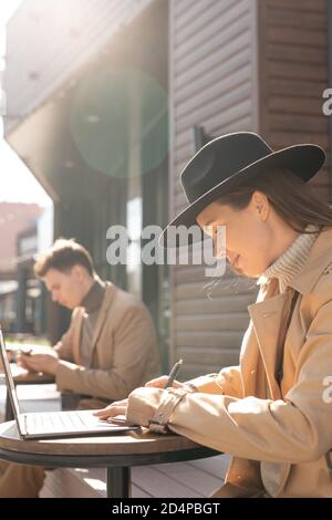 Side view of young elegant female student in hat and trenchcoat making notes Stock Photo