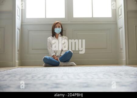 young asian woman in quarantine at home wearing facial mask sitting on floor legs crossed Stock Photo