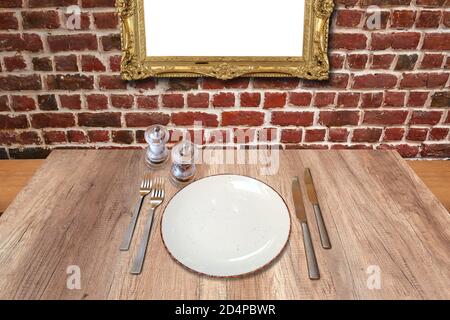 Rustic wooden table with a set of silverware and empty plate against golden frame on red brick wall Stock Photo