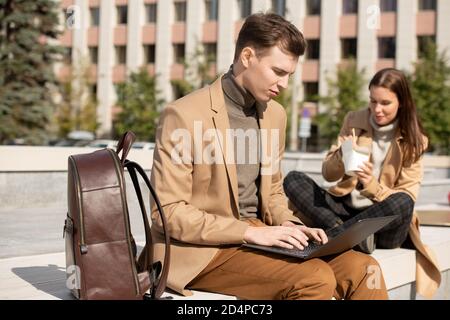 Young serious man using laptop while his girlfriend eating chinese wok Stock Photo