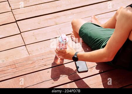 View of young sportsman in green shorts taking bottle of water from wooden bench Stock Photo